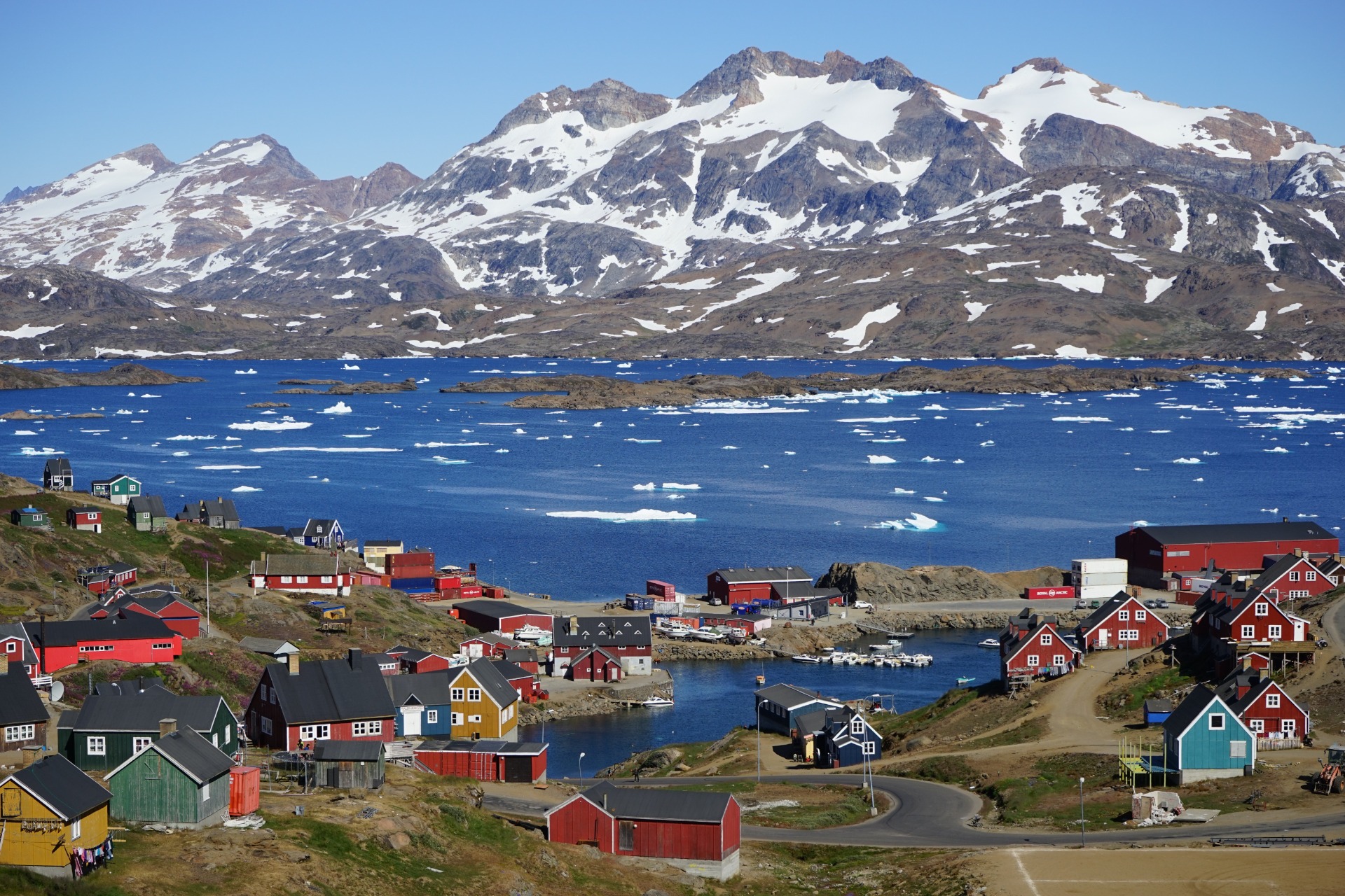 Coastal Greenland village with red houses, icy blue sea, floating icebergs, and snowcapped mountains under clear sky.