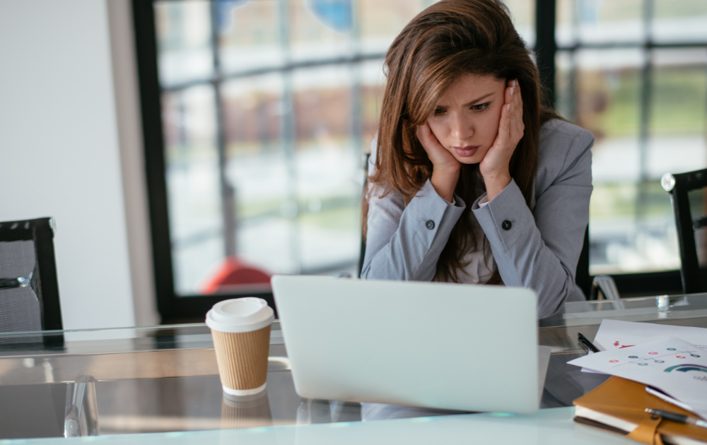 Tired woman in office, resting her head on hands beside laptop and coffee cup.