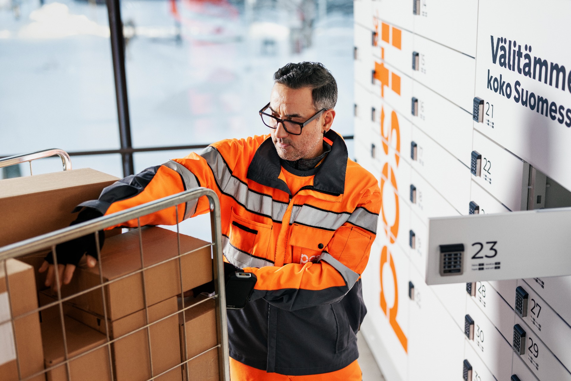 Warehouse worker in orange safety jacket scans packages near lockers with orange numbers and Finnish text. Posti Group, posti, post