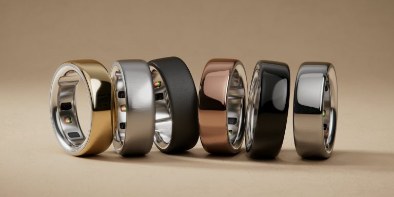 Row of metallic rings in gold, silver, black, and bronze with reflective surfaces on beige background. Oura ring, Oura rings, Oura-sormus, Oura sormus